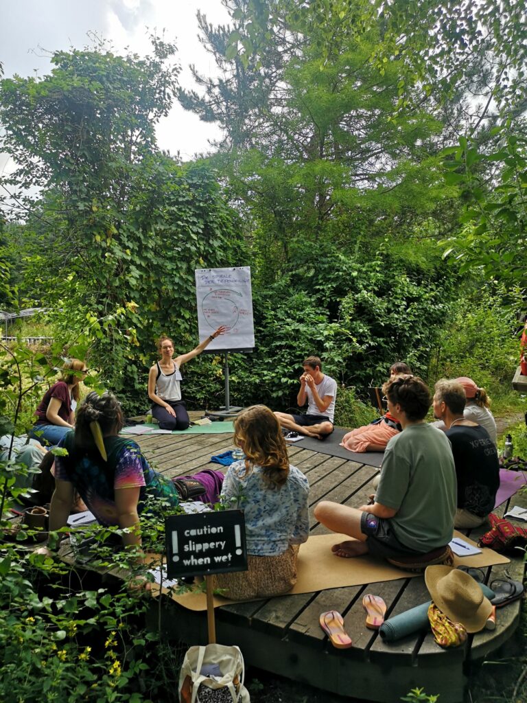 Bild von Menschen die auf einer Holzplattform im Grünen sitzen und einem Vortrag zuhören