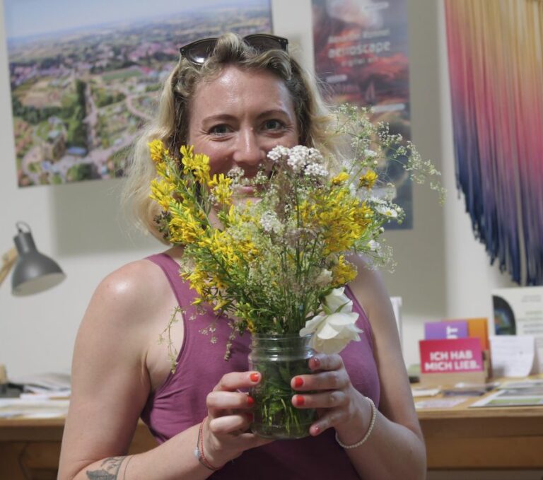 Eine Person lächelt in die Kamera. Ein frisch gepflückter Strauß mit Blumen verdeckt die Hälfte ihres Gesichts